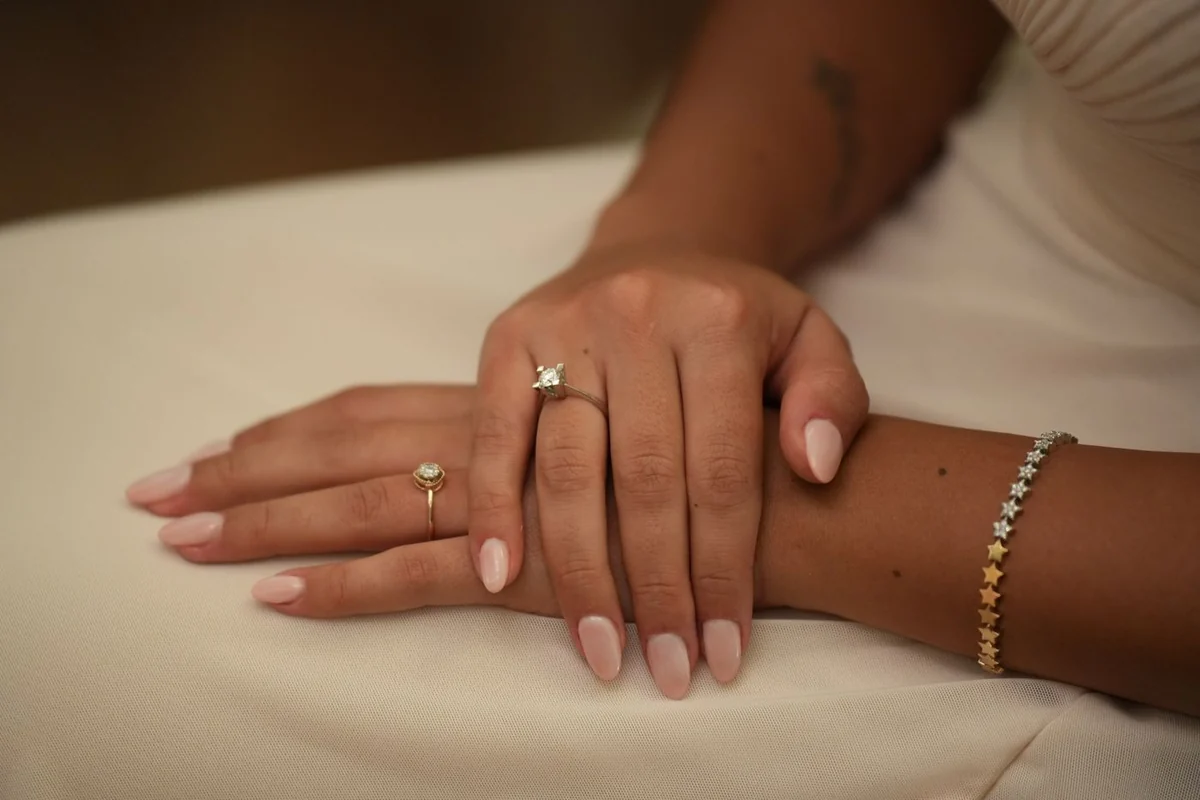 Soft pink manicured nails and delicate gold jewelry against warm ivory fabric